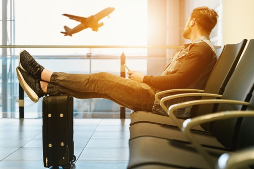 Man sitting in an airport with his legs resting on his carry on, looking out the window at a plane.