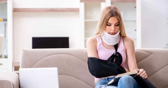 A woman wearing a neck brace and a sling over her right arm sitting on a couch