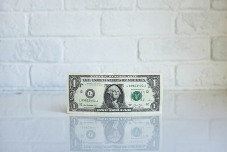 A one-dollar bill upright on a white counter top in front of a white brick wall.