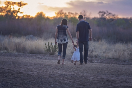 Parents walk outdoors while holding daughter's hand.