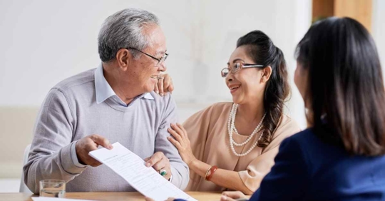 A man and woman smiling as they work with another woman. The man is holding paperwork, and both he and his wife are smiling at each other