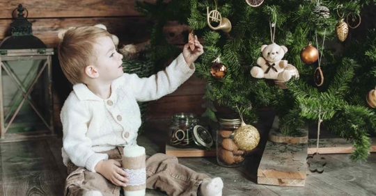 A small child sitting at the base of a holiday tree. The young child is holding an ornament and staring at it with wonder.