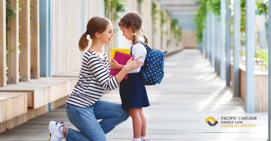 A young mom kneeling in front of her daughter, who is wearing a backpack and carrying books.