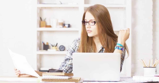 A young woman sitting at her computer. She is reading over a piece of paper