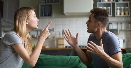 A man and woman sitting on a couch arguing with each other. The woman is pointing at the man, and the man appears defensive.