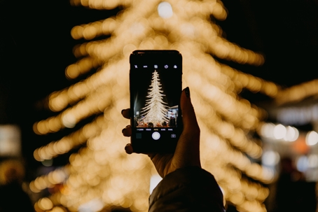 A person holds up a phone to take a picture of a lit Christmas tree.