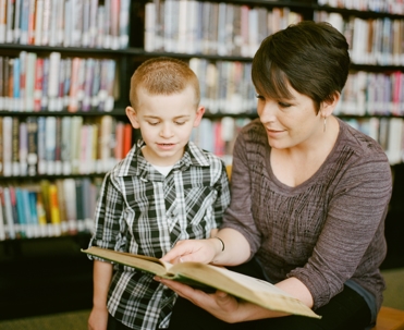 Woman crouches down to help little boy read in library.