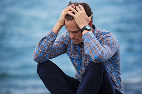 A man sits in front of a body of water with his head in his hands.