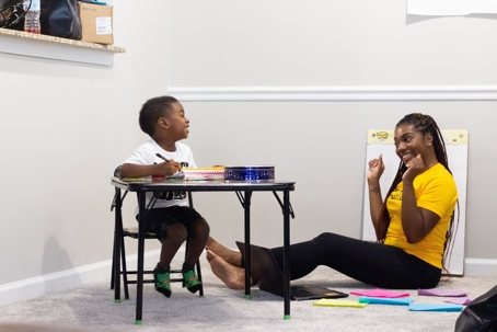 Woman sits on floor teaching young child sitting at desk.