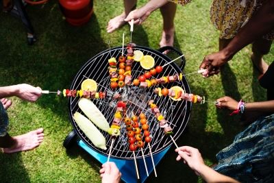 A top-down view of six people holding kebab skewers over a round BBQ grill on a grassy lawn.