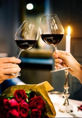 Two people clink wine glasses at dinner over a bouquet of roses in front of a tall candle.