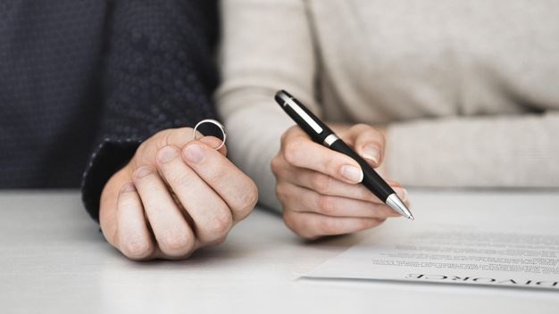 Hand holding a ring next to a hand holding a pen hovering over a paper.